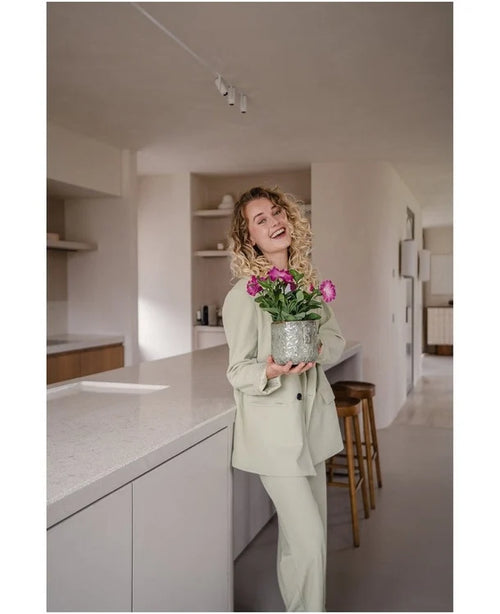 Een glimlachende vrouw met krullend haar, gekleed in een lichtgekleurd pak, staat in een moderne, minimalistische keuken met in haar handen een Kunstbloem Petunia UV 25 cm donkerroze met levendig roze bloemen.