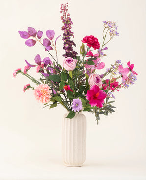 Een vaas met witte structuur bevat het Zijden kunstbloemen boeket Purple Dawn in roze, paarse en lavendeltinten met groen gebladerte, tegen een lichte, neutrale achtergrond.
