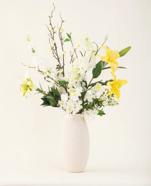 Een vaas met witte structuur toont de elegantie van het Zijden kunstbloemen boeket Sunkissed Ivory, met gele lelies, witte bloemen en groene bladeren tegen een effen lichte achtergrond.