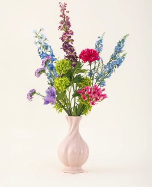 Het Zijden kunstbloemen boeket Blossom Breeze, gerangschikt in een roze vaas van keramiek, heeft levendige blauwe, paarse, rode en groene bloemen - een onderhoudsvrij bloemenboeket tegen een effen lichte achtergrond.
