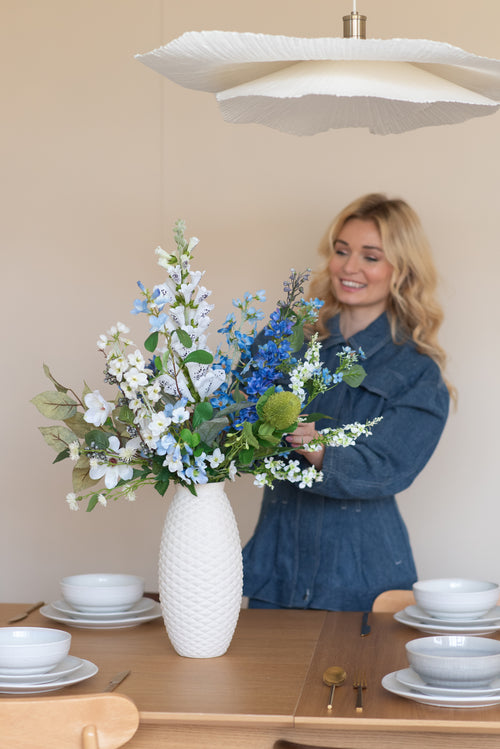 Een glimlachende vrouw in een blauwe spijkerjurk schikt het Zijden kunstbloemen boeket Cool Breeze in een gestructureerde witte vaas op een eettafel gedekt met wit servies en goud bestek.