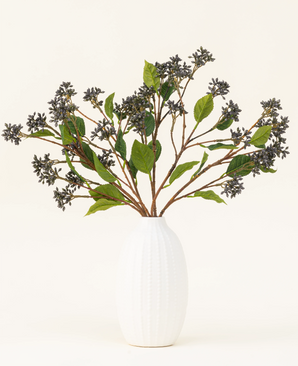Een Zijden kunstbloemen boeket Japanse Berry Staalblauw met lange stelen, donkerpaarse bessen en staalbladeren staat in een vaas met witte textuur tegen een effen lichte achtergrond.