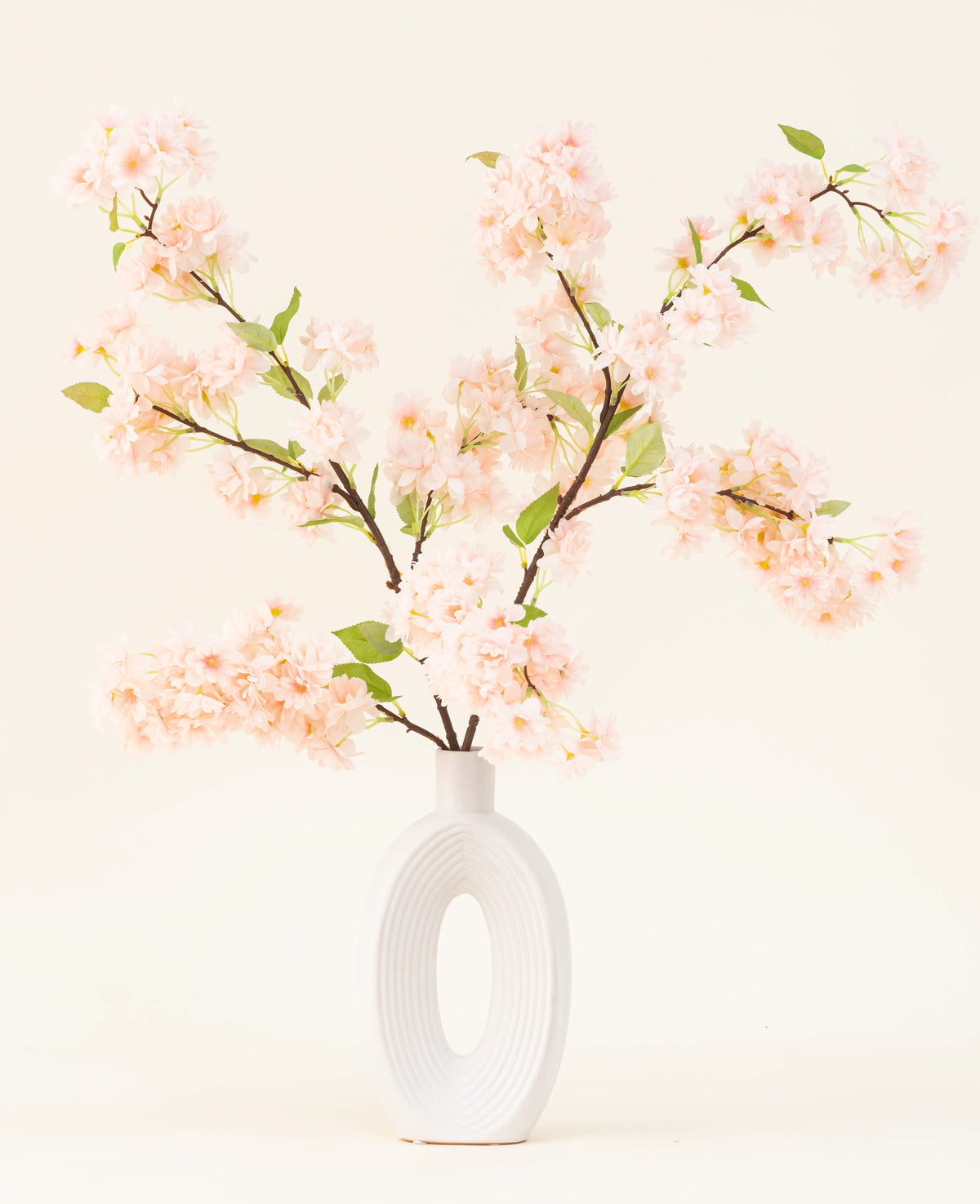 Een witte keramieken vaas met een brede ovale opening toont verschillende groenbladige Zijden kunstbloemen boeket Kersenbloesem lichtroze takken tegen een eenvoudige lichte achtergrond.