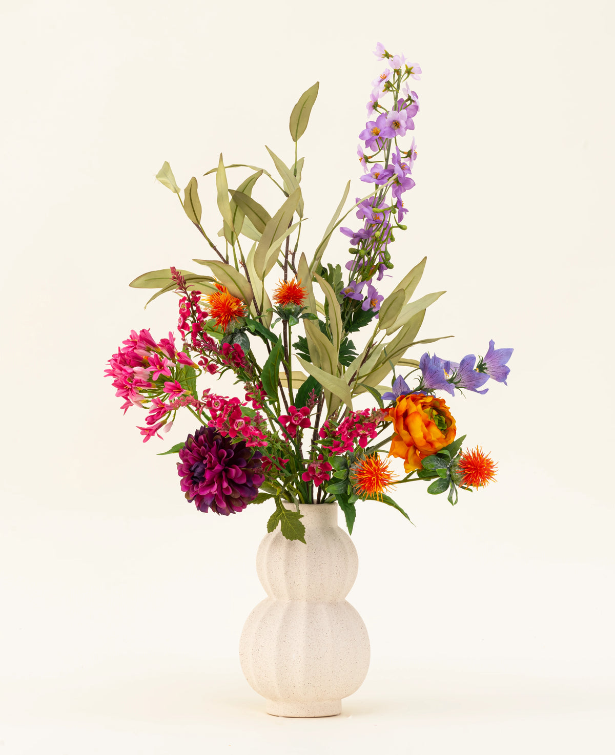 Een witte, gestructureerde vaas toont het Zijden kunstbloemen boeket Colourful Svenski, met levendige paarse, oranje, roze en blauwe kunstbloemen met groene bladeren die elke ruimte opfleuren tegen een eenvoudige lichte achtergrond.