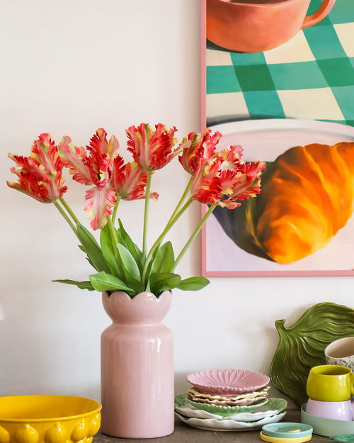 Op een tafel met kleurrijk servies staat een roze vaas met daarin de Kunstbloem Franse Parkiet Tulp 76 cm rood/geel, terwijl op de achtergrond gedeeltelijk een ingelijst schilderij van een croissant en een kopje te zien is.