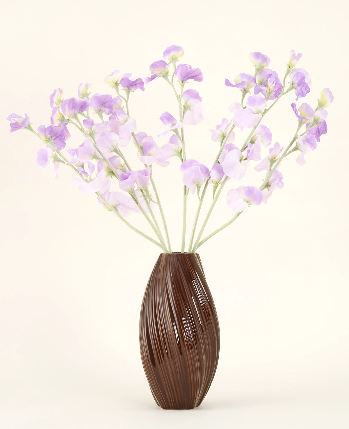 Een bruine vaas met textuur bevat het Zijden kunstbloemen boeket Erwt paars-lichtpaarse zijden bloemen met groenachtige stelen tegen een effen, lichte achtergrond.