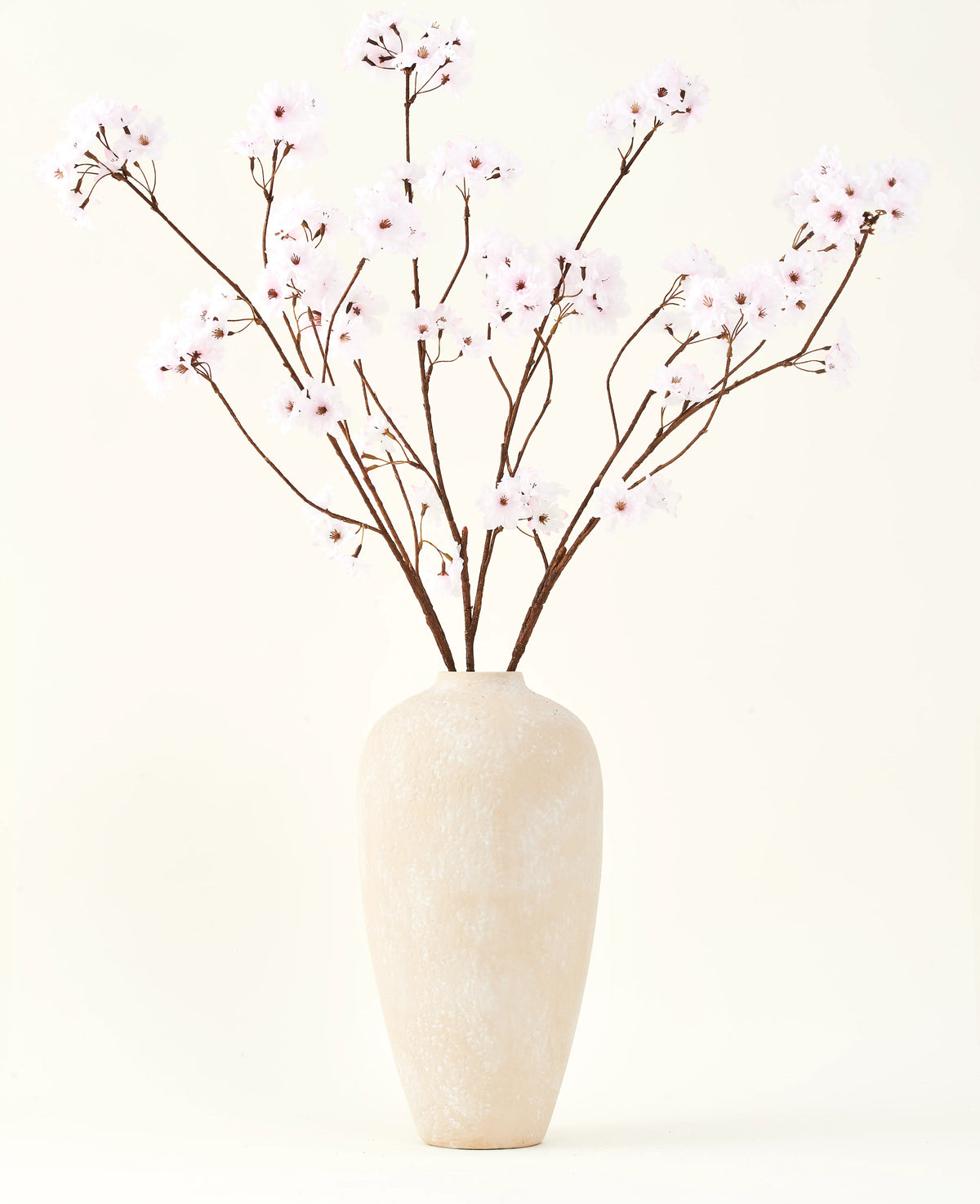 De Zijden kunstbloemen boeket Kersenbloesem lichtroze 114 cm staat in een hoge vaas van beige keramiek met lange, dunne takken en delicate lichtroze bloemen, waardoor een sereen kunstbloemen accent ontstaat tegen een effen achtergrond.