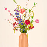 Een April Joy-boeket van kleurrijke zijden bloemen, waaronder roze tulpen en felblauwe takjes, geschikt in een perzikkleurige vaas.