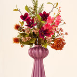 Zijden kunstbloemenboeket Cinnamon Ruby in tinten rood, roze en groen, gepresenteerd in een geribbelde, paarse vaas.