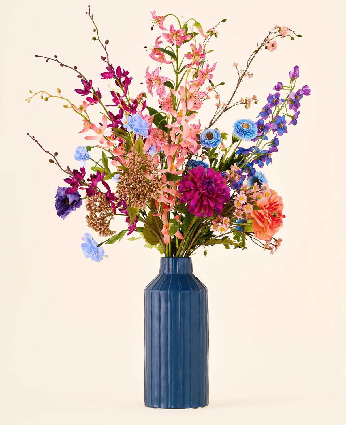 Een groot boeket kleurrijke kunstbloemen van Zijden, het Mikaya-boeket, met onder meer lelies, dahlia’s en orchideeën, in een hoge, geribbelde blauwe vaas.