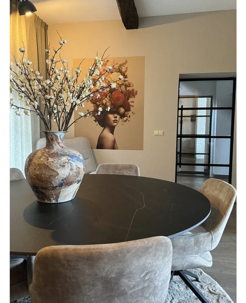 Een moderne eethoek met een ronde zwarte tafel, beige stoelen en een grote keramieken vaas met Kunst Japanse Kersenbloesem 94 cm wit. Een muurschildering van een vrouw met bloemen in haar haar siert de achtergrond.