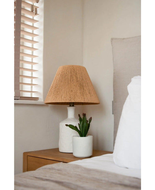 Een nachtkastje met een witte lamp met een geweven beige kap en de Kunst vetplant 20 cm staat naast een bed met wit en beige beddengoed, bij een raam met houten jaloezieën.
