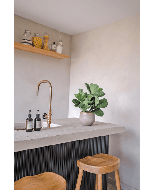 Een modern aanrecht met een messing kraan, twee zeepdispensers, de Kunstplant Tabaksplant Fiddle Leaf UV 45 cm potplant en twee houten krukjes. Een zwevende plank met potten en flessen is gemonteerd op een lichtgekleurde muur.