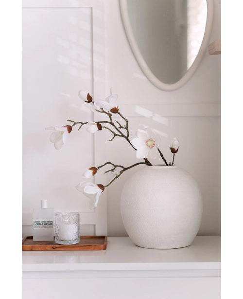 Een Kunstmagnoliatak Real Touch Wit 98cm staat in een ronde witte vaas op een wit oppervlak naast een brandende glazen kaars en een klein flesje op een houten dienblad, in een lichte, minimale kamer met een ronde spiegel erboven.
