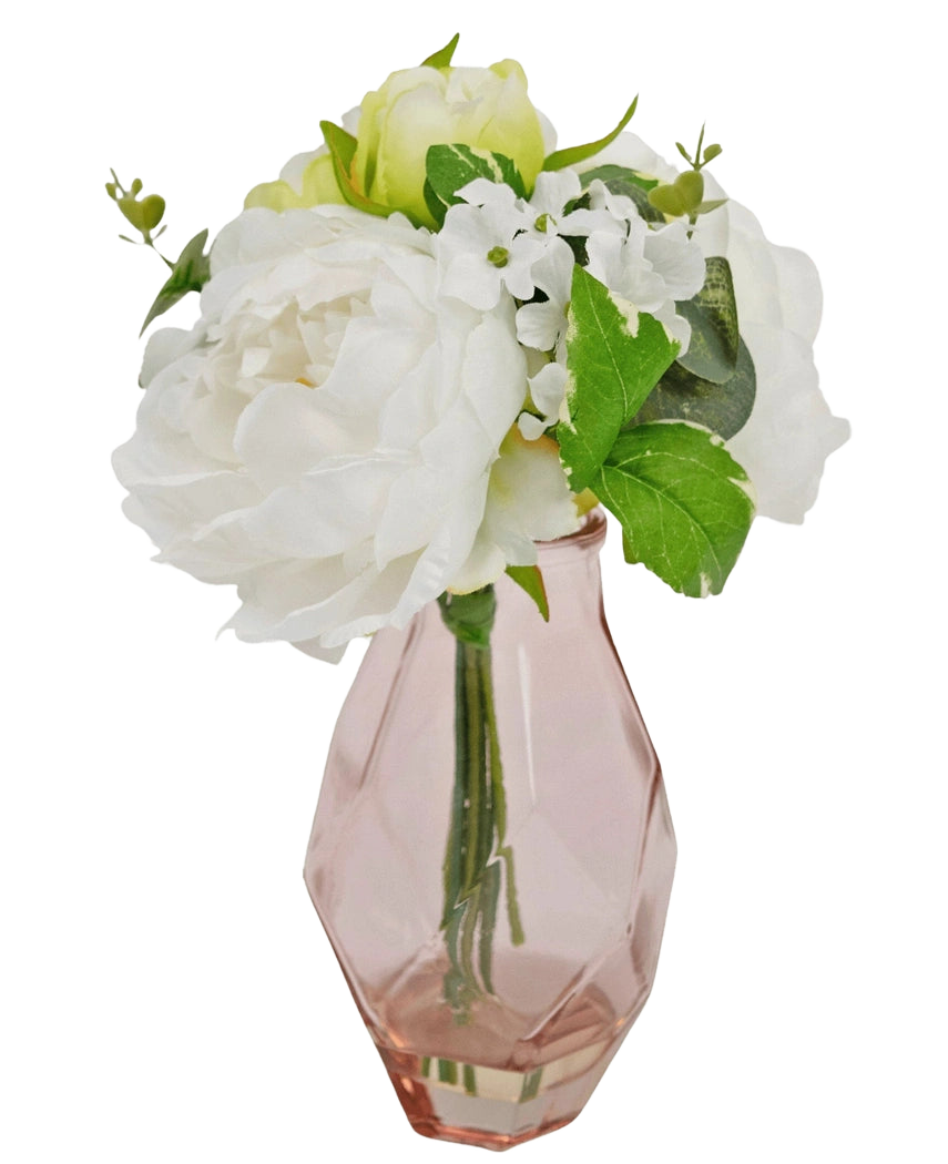Het Kunstboeket in glas 25 cm heeft een klein boeket van witte bloemen met groene bladeren, gerangschikt in een facetgeslepen, lichtroze glazen vaas.