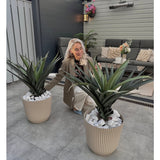 Een vrouw met lang blond haar knielt en glimlacht tussen twee Kunstplant Agava Pandora UV 85 cm potplanten op een betegelde patio, met buitenmeubilair en bloemen aan de muur op de achtergrond.