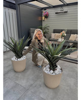 Een vrouw met lang blond haar knielt tussen twee Kunstplant Agava Pandora 85 cm UV met stekelige bladeren op een grijs betegelde patio. Achter haar is een zithoek met kussens, een muur en hangende bloemenmanden.