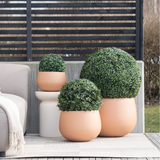 Drie Kunstplant Buxus romeinse stijl UV D30cm ronde terracotta plantenbakken met afgezette groene struiken staan op een patio naast een beige buitenbank en een klein wit tafeltje, met daarachter een privacyscherm met latten.
