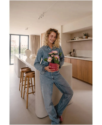 Een vrouw in spijkerbroek staat in een moderne keuken, glimlachend en met de Kunstplant Gerbera UV 28 cm roze in haar handen – een kleine roze bloempot met roze en witte bloemen. Op de achtergrond zijn twee houten krukken en een groot raam te zien.