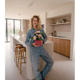 Een persoon in een spijkeroutfit staat in een moderne keuken en glimlacht naar de camera terwijl hij of zij de Kunstplant Gerbera UV 28 cm roze vasthoudt. Rond het keukeneiland staan houten krukken opgesteld en op de achtergrond zijn decoratieve planken te zien.