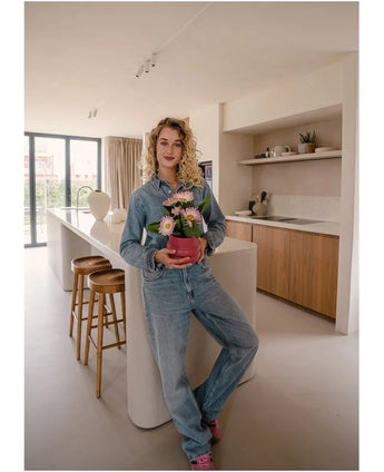 Een persoon in een spijkeroutfit staat in een moderne keuken en glimlacht naar de camera terwijl hij of zij de Kunstplant Gerbera UV 28 cm roze vasthoudt. Rond het keukeneiland staan houten krukken opgesteld en op de achtergrond zijn decoratieve planken te zien.