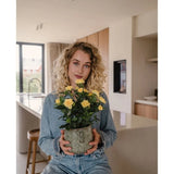 Een vrouw met krullend blond haar, gekleed in spijkerkleding, zit in een moderne keuken met de Kunstplant Mini Rose UV 35 cm geel in haar handen en kijkt recht in de camera.