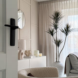 Een moderne, minimalistische woonkamer met een beige fauteuil met textuur, Kunstplant Dracaena Fragnans 180 cm, wit dressoir met decor, ronde spiegel en transparante gordijnen die zacht daglicht filteren door het raam.