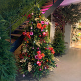 De Kunstkerstboom "Weideman" PE/PVC - Smart LED staat binnen bij een trap, versierd met roze en rode bloemen, gouden bladeren en roze ornamenten, omgeven door groen op een gepolijste betonnen vloer voor een stijlvolle kerstversiering.