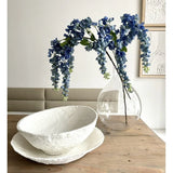 Een bloemstuk van Kunst Blauweregen van 115 cm staat in een doorzichtige vaas op een houten tafel, naast twee witte schalen met een structuur, terwijl de muur erachter versierd is met abstracte kunst.