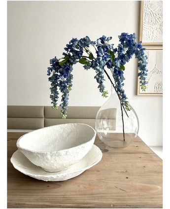 Een bloemstuk van Kunst Blauweregen van 115 cm staat in een doorzichtige vaas op een houten tafel, naast twee witte schalen met een structuur, terwijl de muur erachter versierd is met abstracte kunst.