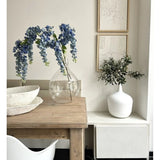 Op een houten tafel staat een doorzichtige vaas met daarin de Kunst Blauweregen van 115 cm, naast een witte schaal. Vlakbij staat een witte vaas met groen op een witte standaard, onder twee ingelijste kunstwerken met een structuur op een lichte muur.