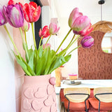 Een vaas met roze hartmotieven houdt Kunstbloem Franse Tulp 60 cm paars op een eettafel met stoelen met geweven rugleuning. Op de achtergrond dragen een ronde spiegel en een muur met bruin patroon bij aan het gezellige, moderne interieur.