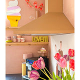 Een pastelroze keuken met Kunstbloem Franse Tulp 60 cm paars op de voorgrond, een geel LOVETTI bord op de plank, roze mokken, muurkunst van een ijshoorntje en een minimalistische muurschildering van twee mensen op de muur.