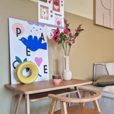 Een klein houten bureau met een stoel is versierd met Kunstbloem Catnip 75 cm roze, een mok, een gele ringsculptuur en kleurrijke kunstprints aan de muur erboven. Rechts is een bank gedeeltelijk zichtbaar.