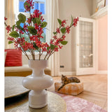 Op een tafel staat een witte vaas met roze Kunsttak-bessen (64 cm) en groene bladeren. Op de achtergrond ligt een kat op een roze kussen in een lichte woonkamer met beige muren en een glazen deur.