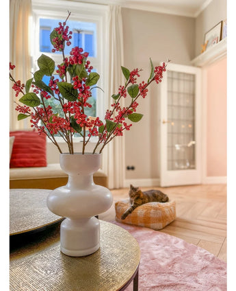 Op een tafel staat een witte vaas met roze Kunsttak-bessen (64 cm) en groene bladeren. Op de achtergrond ligt een kat op een roze kussen in een lichte woonkamer met beige muren en een glazen deur.
