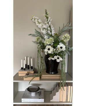 Op een plank tegen een lichte muur staat een zwarte vaas met de Kunstbloem Amaranthus 124 cm groen, naast vijf witte kaarsen, beige boeken en decoratieve voorwerpen in neutrale tinten.