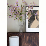In een doorzichtige vaas staan de bloemen van de Kunsttak Daphne Spray 108 cm paars op een donkerhouten oppervlak naast een modern, abstract kunstwerk in een lijst, met daaronder een witte lampenkap met structuur.