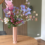 Een hoge roze en wit gestreepte vaas met het zijden kunstbloemen boeket Amethyst Romance staat op een ronde houten tafel in een moderne kamer met groene muren, houten lambrisering en kleine ingelijste kunst.