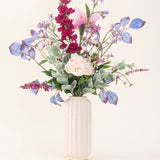 Een vaas met witte structuur toont het "Zijden kunstbloemen boeket Amethyst Romance" met roze pioenrozen, paarse en blauwe bloemen, bordeauxrode bloemen en groen gebladerte tegen een lichte, neutrale achtergrond.