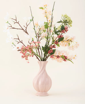 Een roze vaas van keramiek bevat het Zijden kunstbloemen boeket Springtime Poem, met witte, roze en groene bloemen met groene bladeren en bruine stelen, tegen een lichte achtergrond.