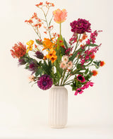 Een witte, geribbelde vaas van keramiek toont het Zijden kunstbloemen boeket Magnus di Lambo - een arrangement van levendige oranje, roze, paarse en gele kunstbloemen met groene bladeren tegen een lichte achtergrond.
