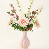 Een roze vaas van keramiek bevat het Zijden kunstbloemen boeket Morning Whisper, met lichtroze dahlia's, witte leeuwenbekjes, groene bladeren en trosjes kleine rode besjes, tegen een effen gebroken witte achtergrond.