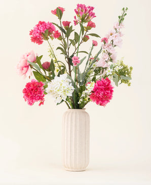 Een vaas met witte textuur toont het Zijden kunstbloemen boeket Blush Breeze - roze, witte en lichtpaarse zijden bloemen met groene bladeren - tegen een effen lichte achtergrond.