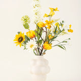 In een witte vaas met structuur staat het Zijden kunstbloemen boeket Sun Glow met gele tulpen, zonnebloemen, madeliefjes en groen blad tegen een effen gebroken witte achtergrond.