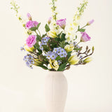 In een crèmekleurige vaas staat een Zijden kunstbloemen boeket Magnolia Glow met paarse, witte en gele bloemen-delphiniums, lisianthus, lelies en vergeet-me-nietjes tegen een effen lichte achtergrond.