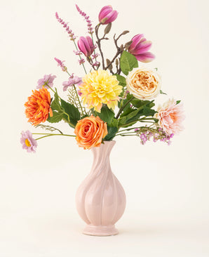 Het Zijden kunstbloemen boeket Sweet Daybreak heeft een pastelroze vaas gevuld met oranje rozen, gele dahlia's, roze magnolia's en lichtpaarse bloesems, prachtig gerangschikt tegen een effen lichte achtergrond.