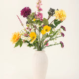 Het Zijden kunstbloemen boeket Daisy Daydream heeft een vaas met witte textuur en een levendige mix van gele, roze, paarse en witte bloemen met groene bladeren, die blijvende schoonheid toevoegt aan elke effen lichte achtergrond.