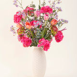 Een vaas met witte structuur en het Zijden kunstbloemen boeket Soft Spring met felroze bloemen, oranje tulpen en paarsblauwe bloesems brengt lente in huis tegen een effen lichte achtergrond.