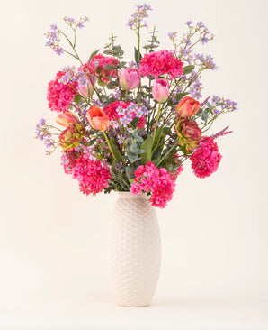 Een vaas met witte structuur en het Zijden kunstbloemen boeket Soft Spring met felroze bloemen, oranje tulpen en paarsblauwe bloesems brengt lente in huis tegen een effen lichte achtergrond.